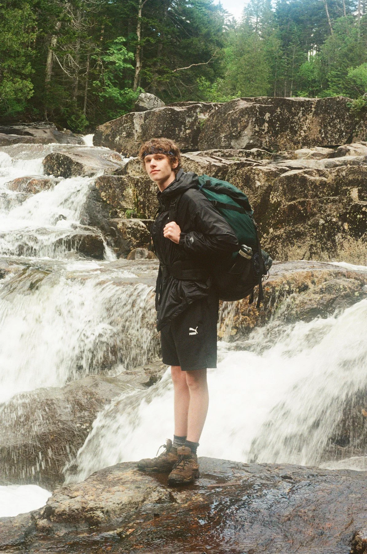 Young man in outdoor gear standing on a wet rock near a flowing waterfall surrounded by greenery.