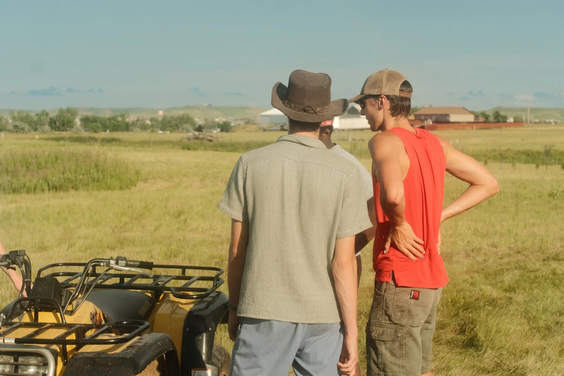 Two men standing in a grassy field talking, one wearing a cowboy hat and grey shirt, the other wearing a cap, red tank top, and shorts, nearby an ATV.