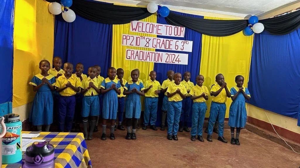 Group of children in yellow and blue school uniforms standing on a stage with a yellow, blue, and black backdrop, celebrating graduation.