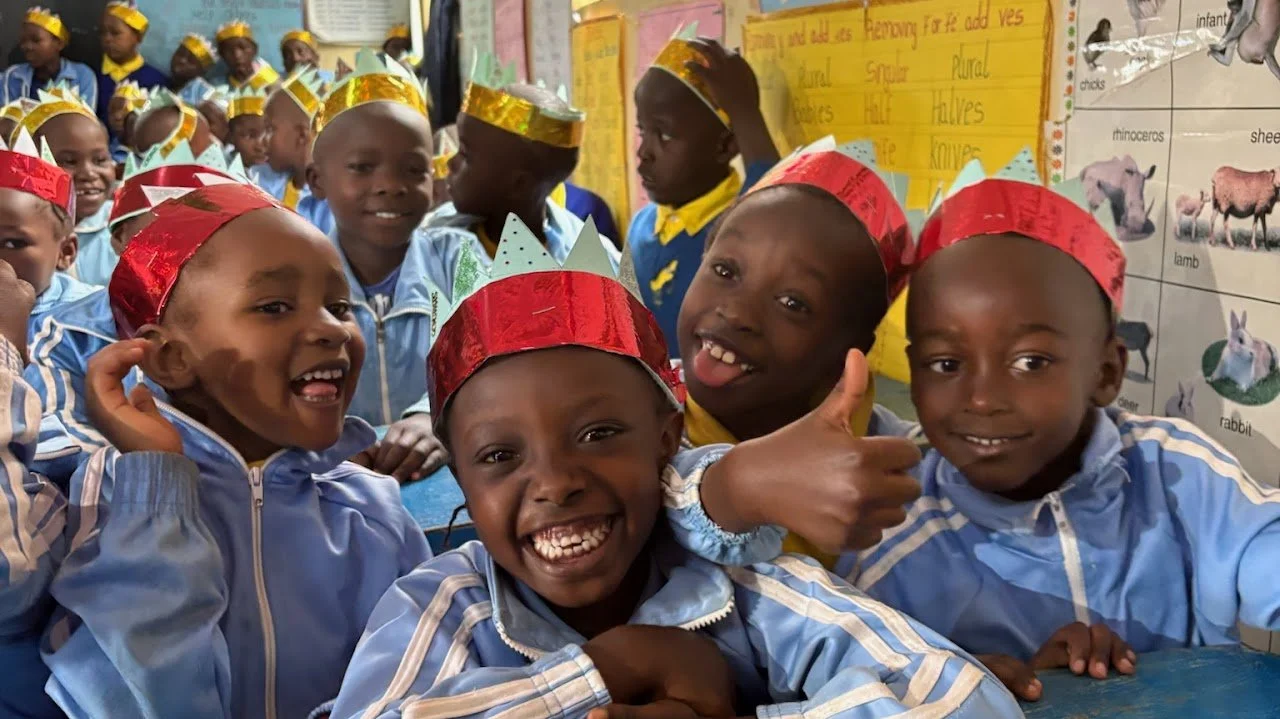 Young children in a classroom wearing colorful paper hats, smiling and celebrating, with educational posters visible in the background.