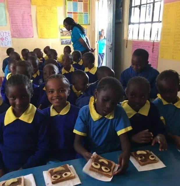 Group of young students in blue and yellow school uniforms inside a classroom, some smiling at the camera, with baked goods on the table in front of them.
