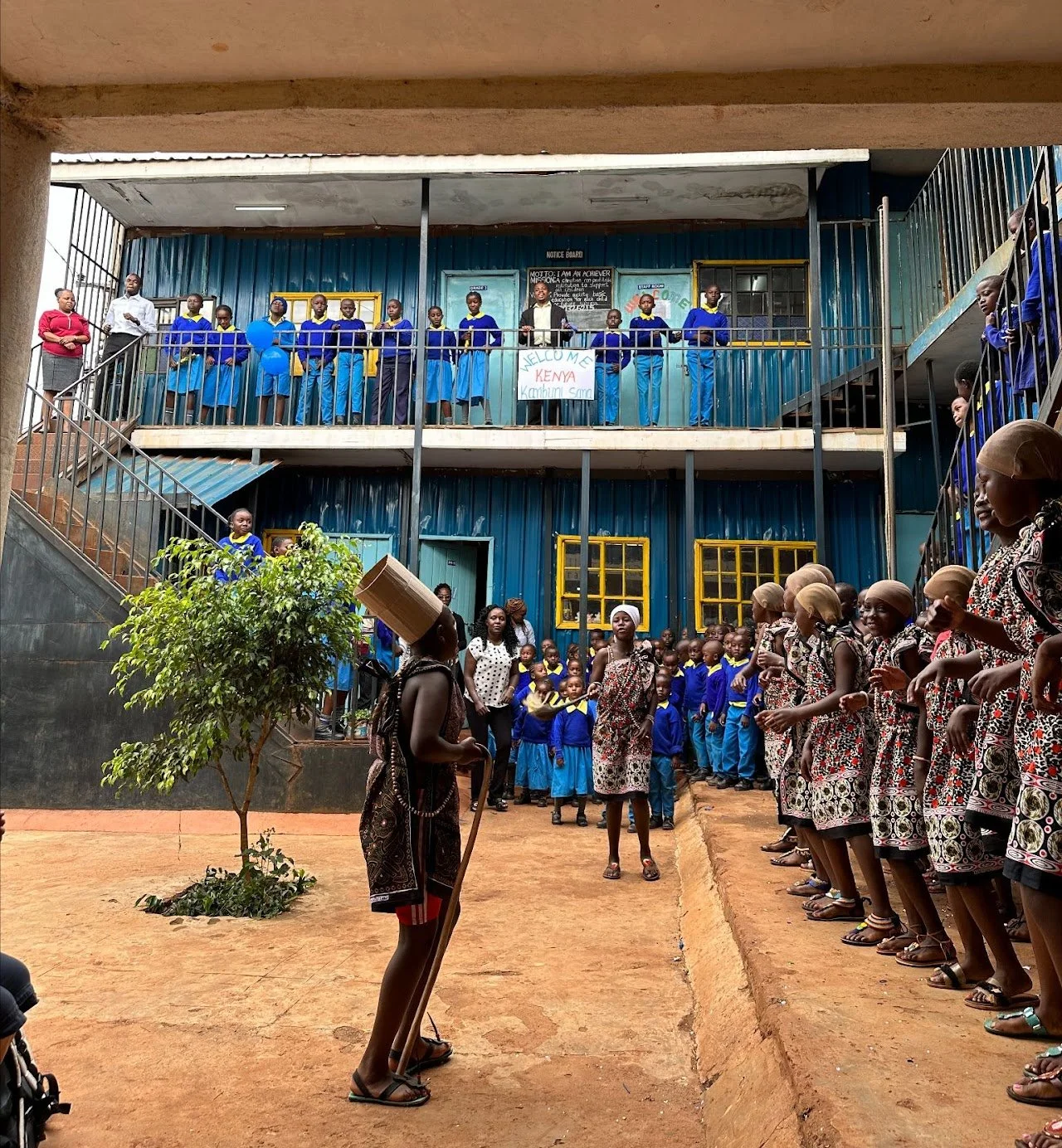 School children in Kenya wearing uniforms, gathered in an outdoor courtyard and on the balcony, participating in a cultural event with traditional dance and music, during the daytime.