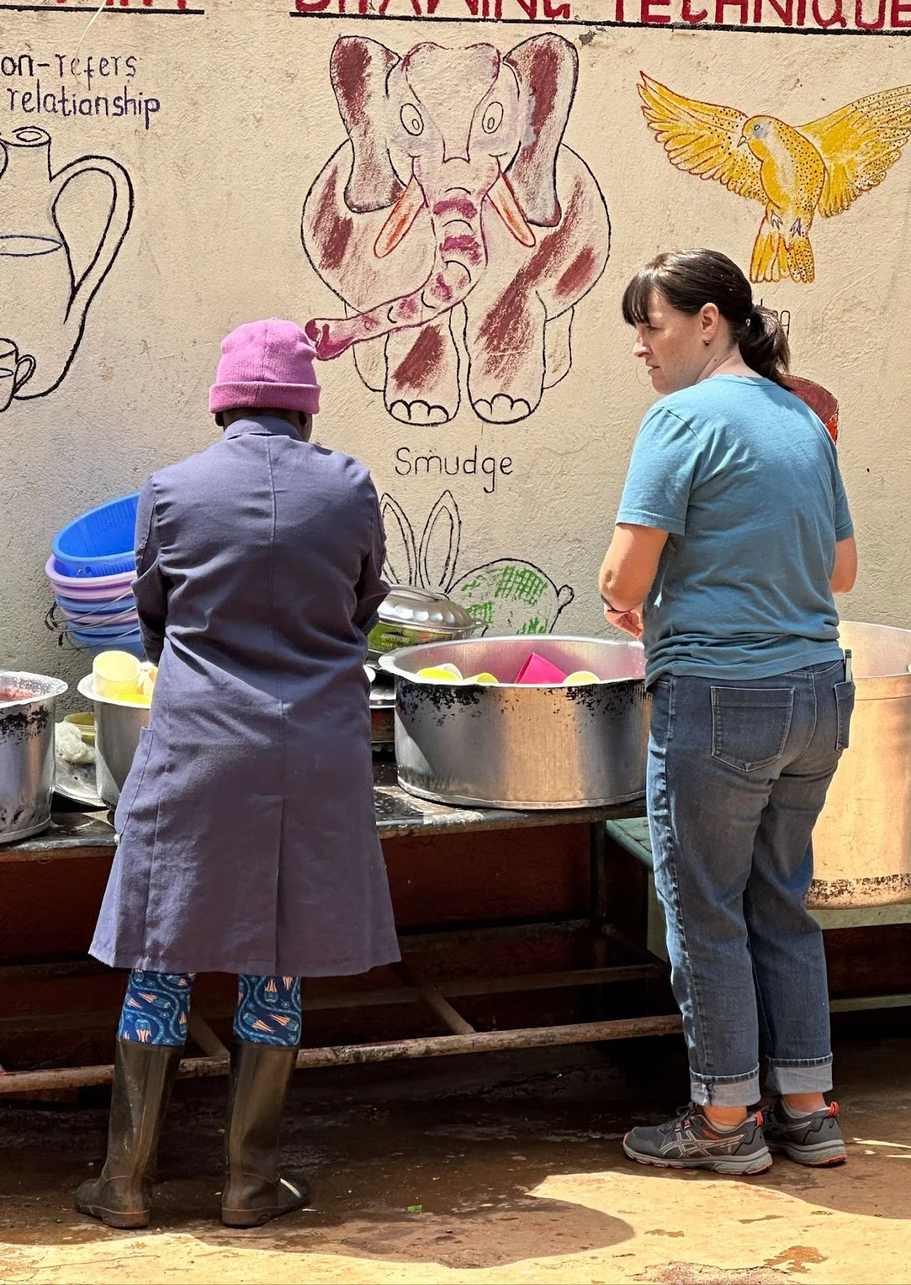 Two women standing outside near a table with large metal bowls, preparing food. One woman wears a pink beanie, gray coat, and rubber boots, while the other wears a blue t-shirt, jeans, and sneakers. Behind them, a wall features a colorful drawing of 