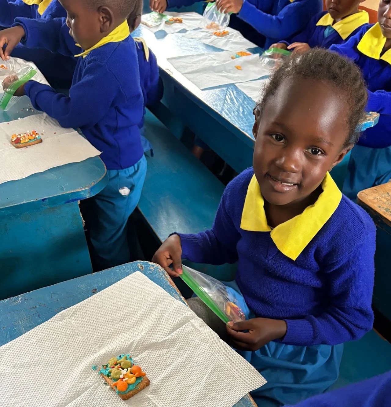 Young girl in blue school uniform with yellow collar smiling while holding a plastic bag, with decorated cookie on table in front of her, children in similar attire at desks in background.