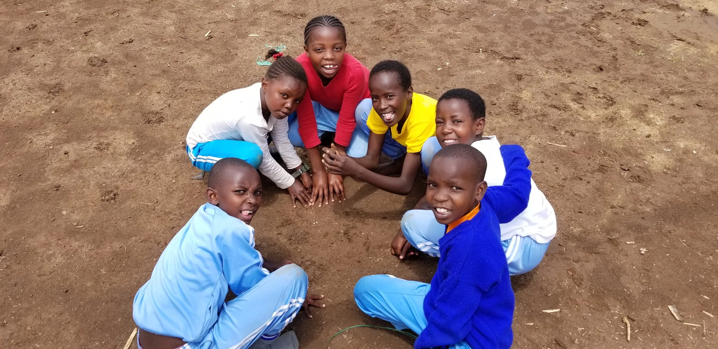 Group of seven young children sitting on dirt ground in a circle, smiling and looking at the camera.