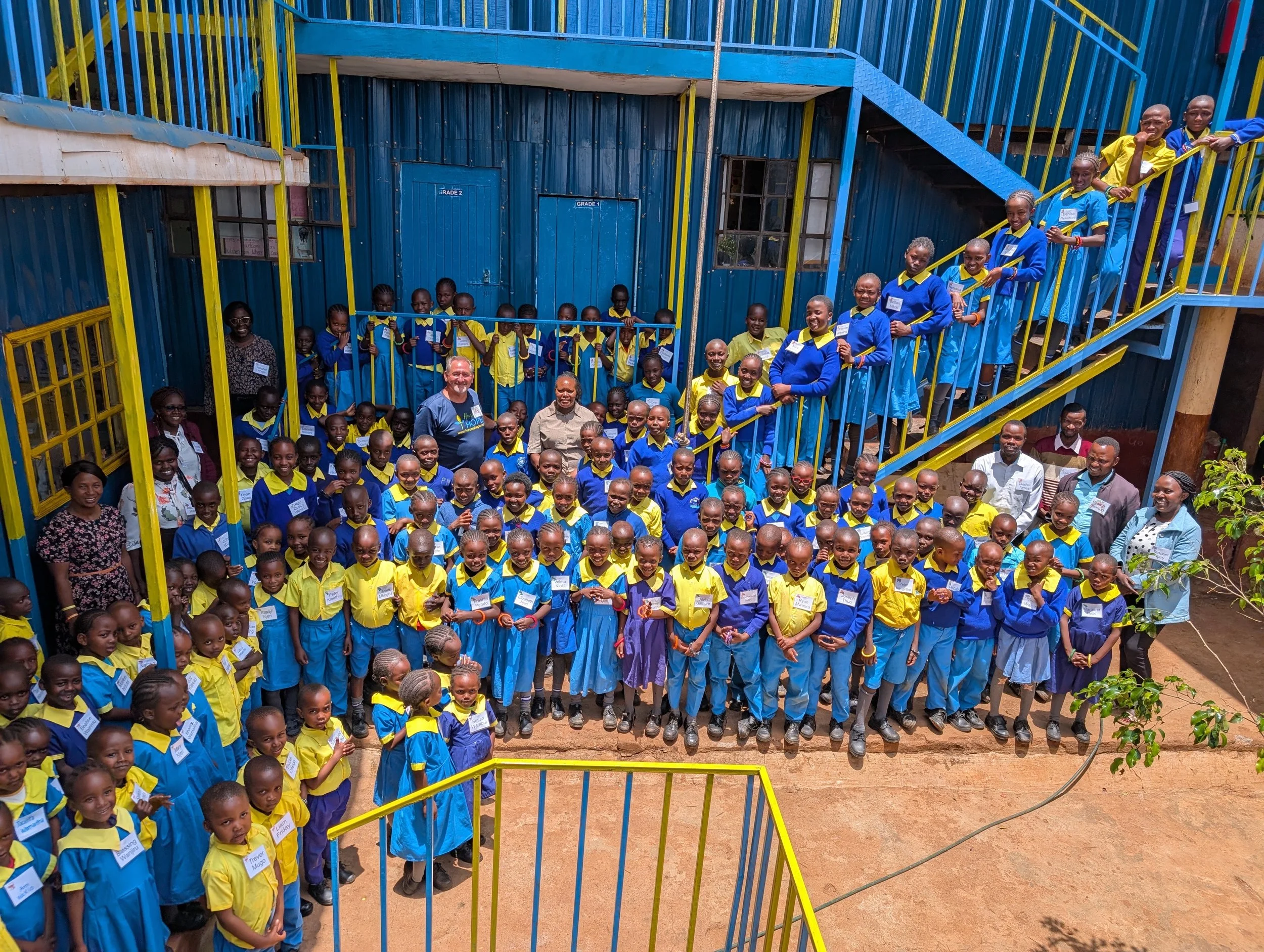 Group of children and adults posing for a photo in a school courtyard with blue and yellow painted buildings and stairs.
