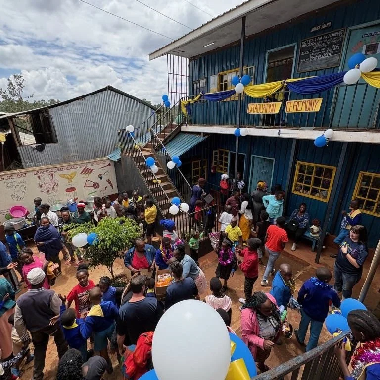 Community graduation ceremony in a schoolyard decorated with yellow, blue, and white balloons, with many children and adults gathered around near a blue building with a staircase and a mural on a nearby wall.