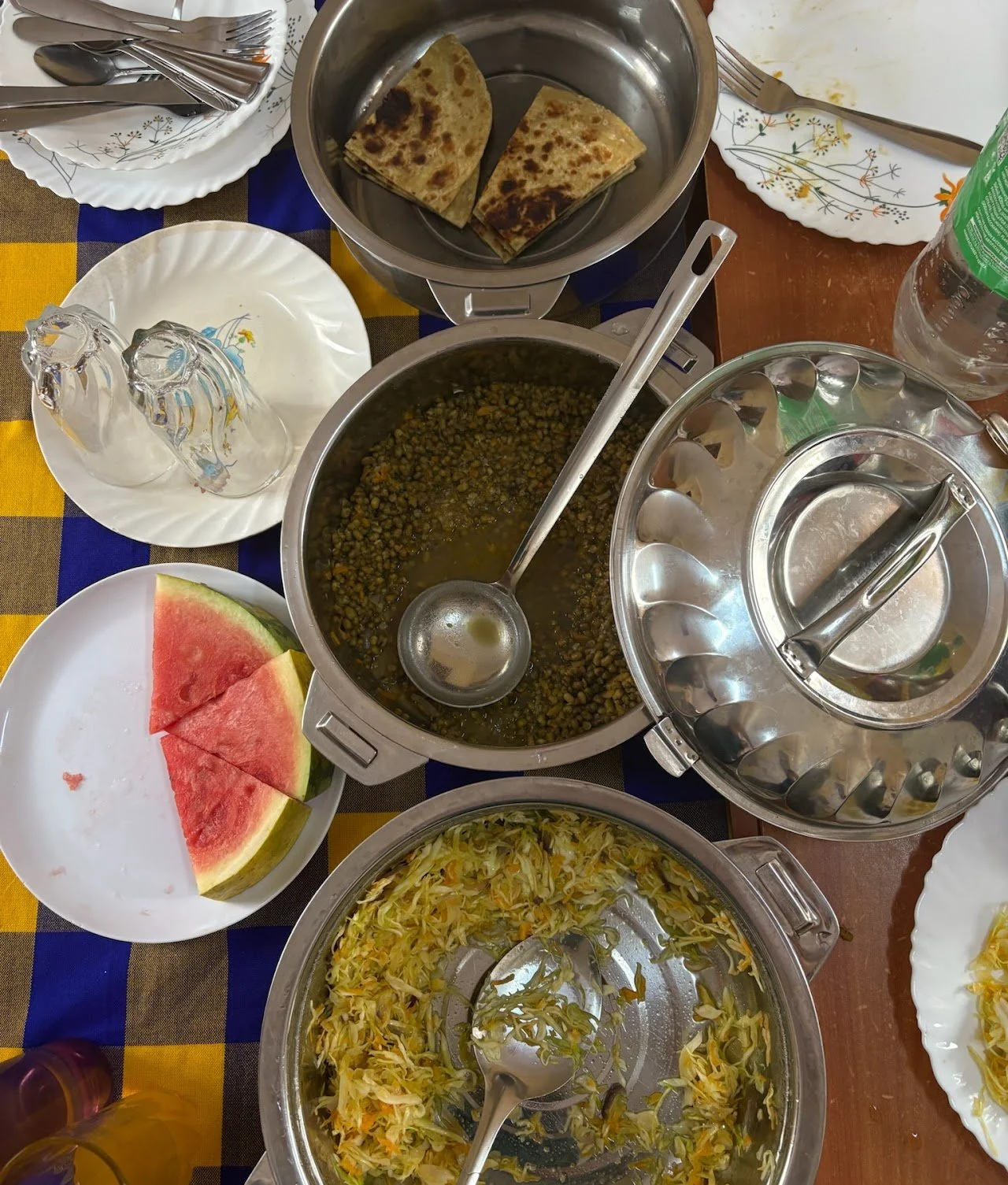 A table set with watermelon slices, various dishes including a lentil and vegetable stew, cooked cabbage, and flatbreads. There are plates, glasses, a water bottle, and utensils arranged on a checkered tablecloth.