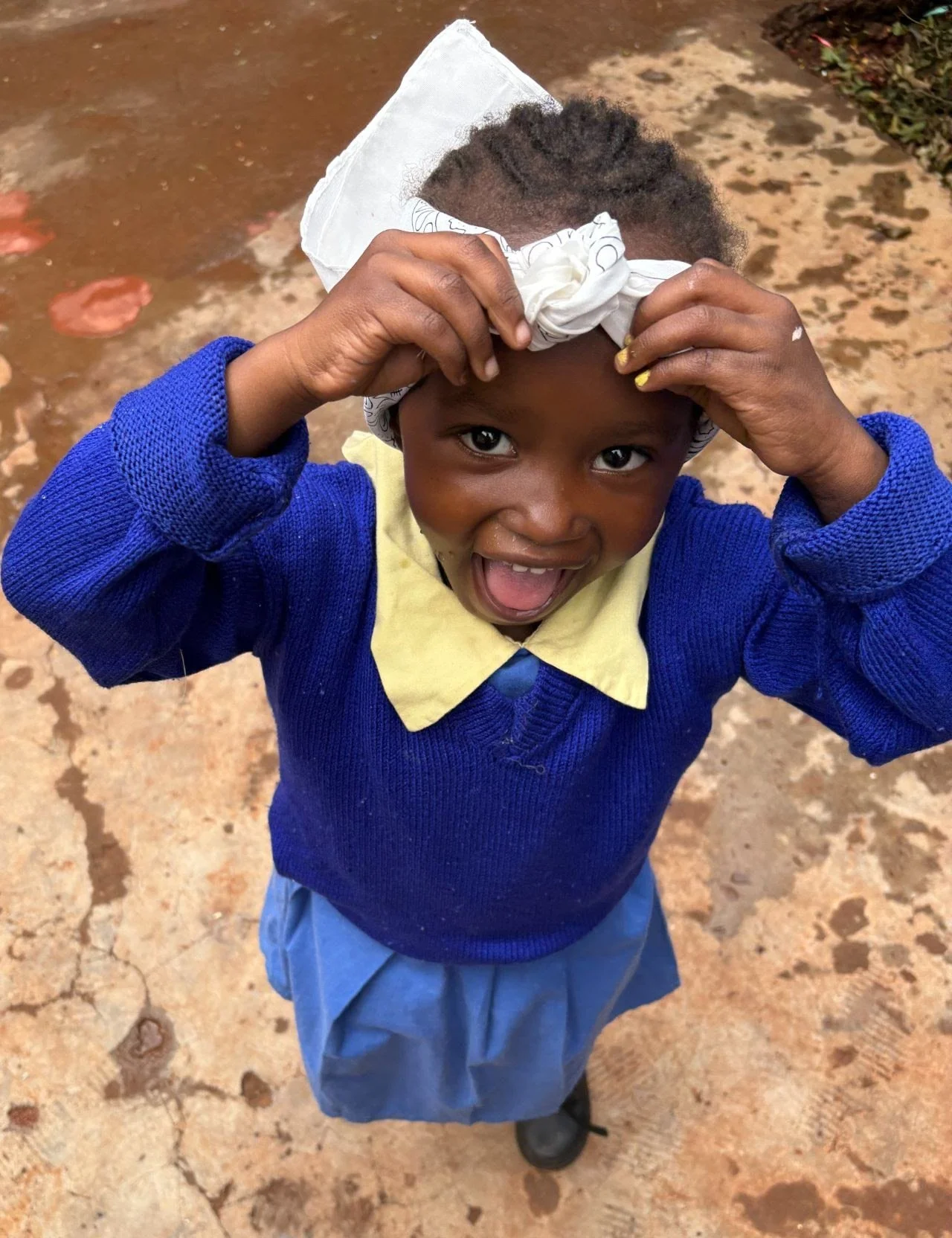 A young girl in school uniform smiling and holding a bandana on her head, looking up at the camera.