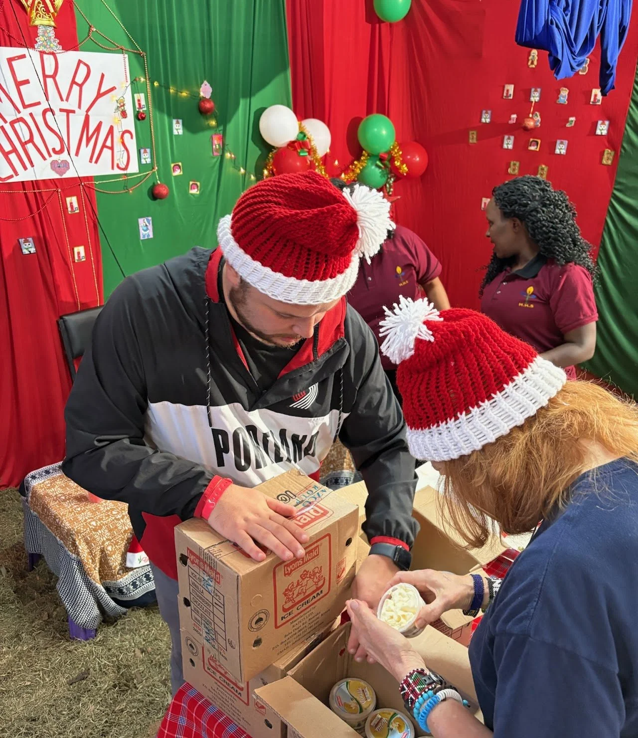 People wearing red and white Christmas hats packaging boxes of ice cream during a holiday charity event with Christmas decorations in the background.