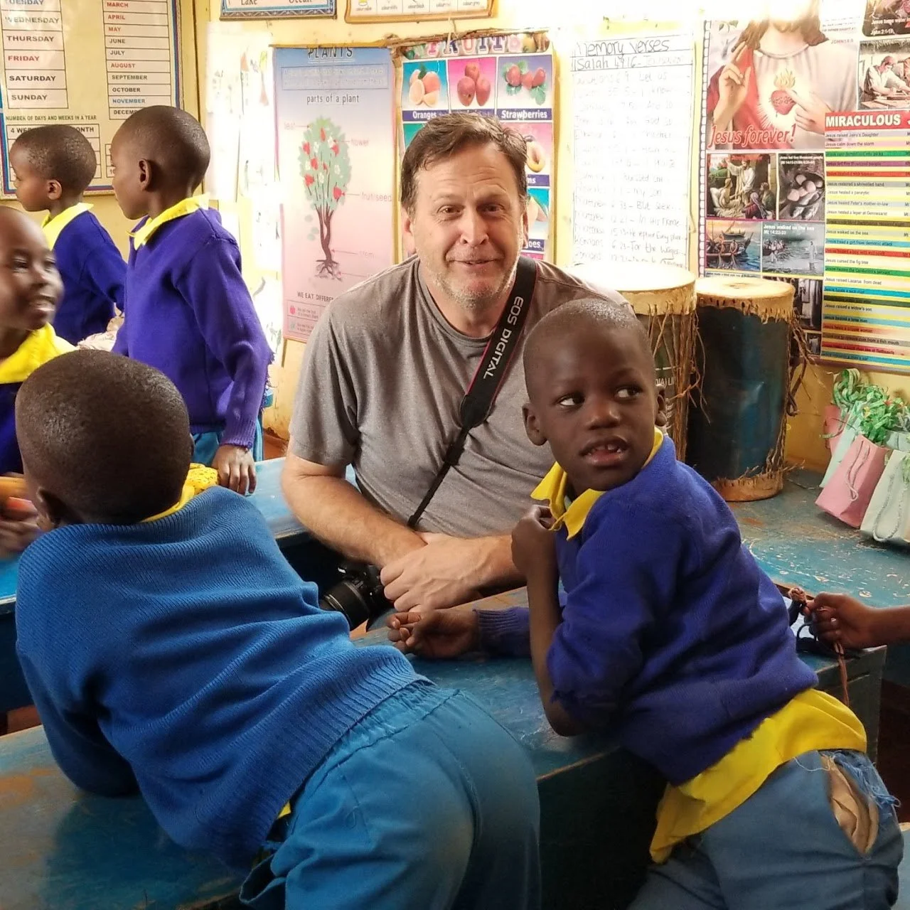 A man with a camera around his neck sitting at a table with young school children wearing blue and yellow uniforms in a classroom, with posters and drums on the wall behind them.
