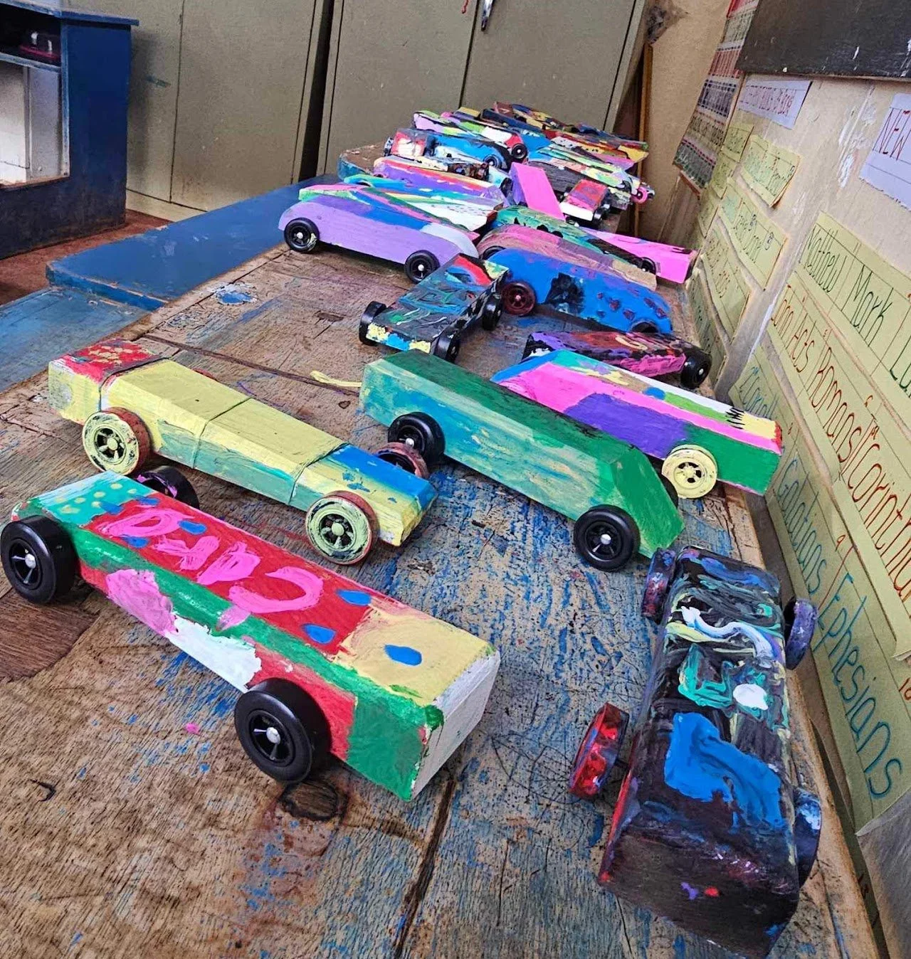 Colorfully painted wooden toy cars on a classroom table with educational posters on the wall.