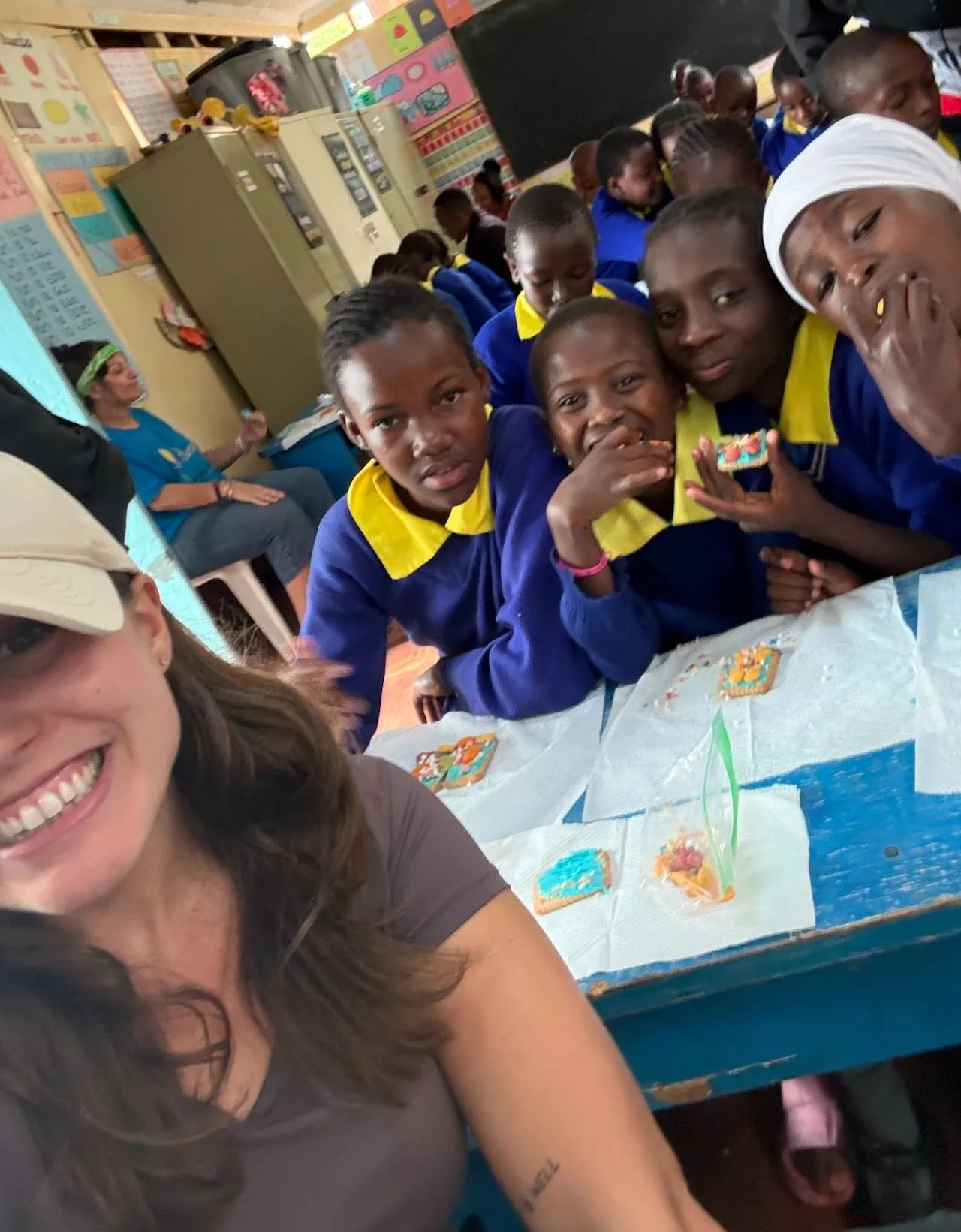 Group of children and an adult taking a selfie in a classroom, children in blue and yellow uniforms, some eating snacks on a table with decorated cookies.