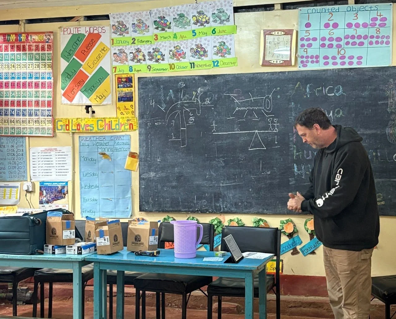 A classroom with a teacher standing in front of a chalkboard. The blackboard has drawings and words, and the wall above has colorful educational posters, including months and a countdown chart. There are tables with boxes of supplies, a purple pitche
