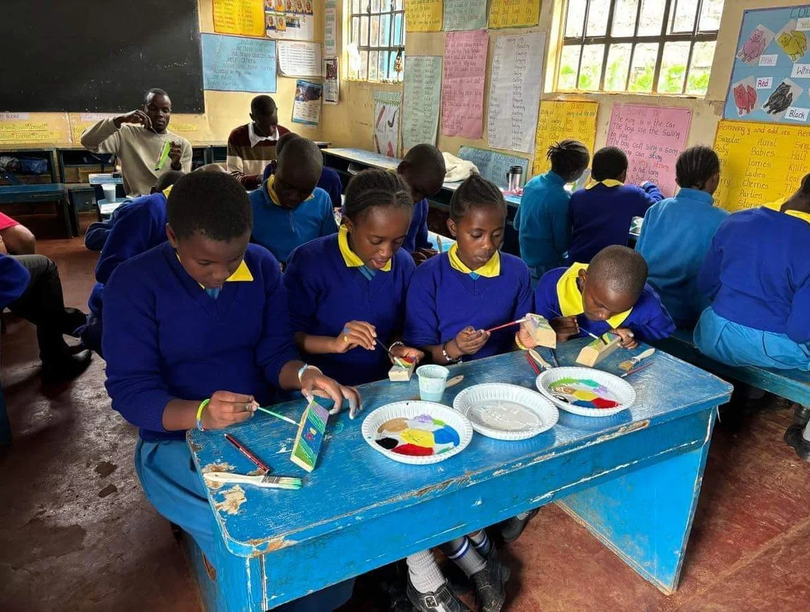 Students in a classroom are painting small objects at a blue desk, with colorful posters on the walls and large windows letting in natural light.
