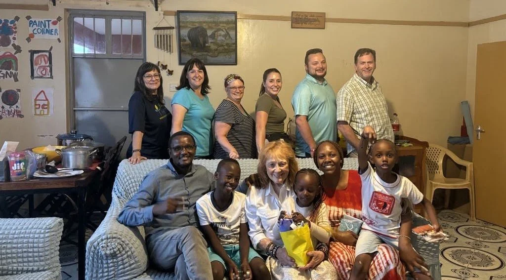 Group of nine people, including children and adults, gathered in a living room for a photo. Some are sitting on the couch, and others are standing behind it. The room has artwork on the walls and some children’s drawings on the door. There are tables