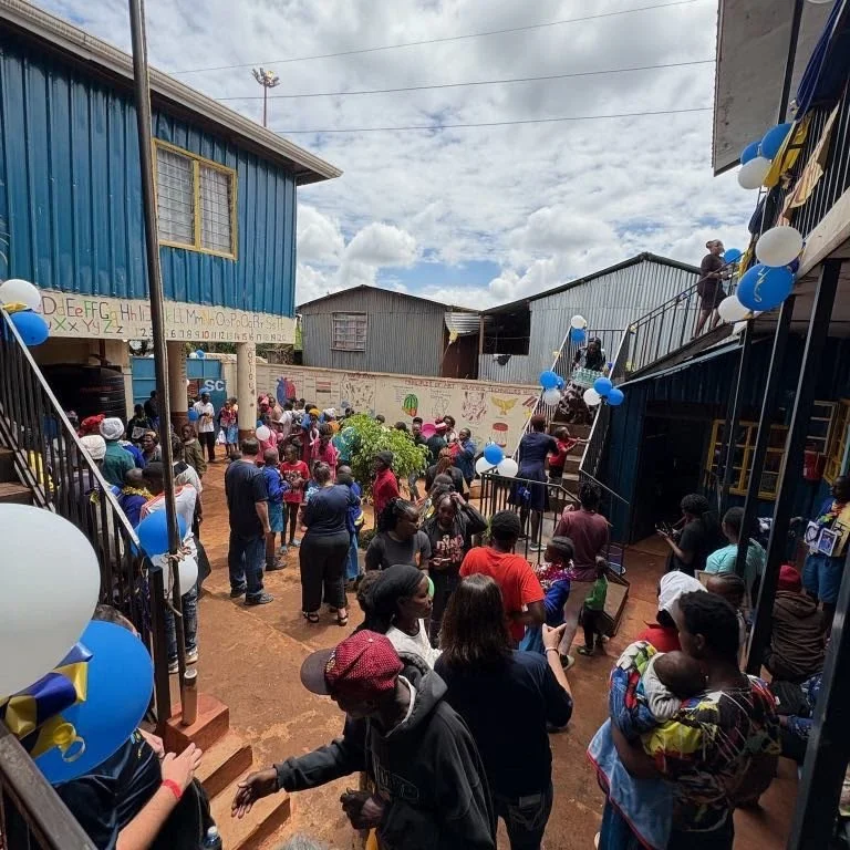 Crowd of people gathered in an outdoor courtyard decorated with blue and white balloons, attending a community event or celebration.