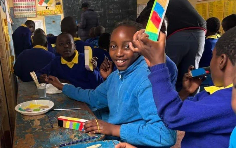 School children in uniforms sitting at desks in a classroom, with one smiling boy showing a colorful object.