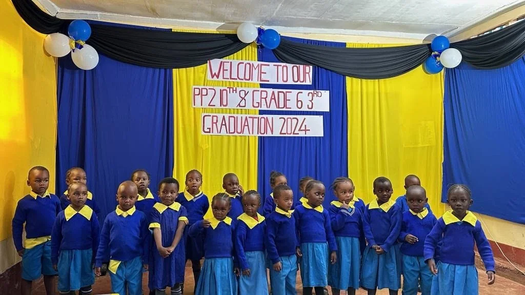 Group of young children in blue and yellow uniforms standing on a stage with a banner that reads 'Welcome to our PP 2 10th & Grade 6 3rd Graduation 2024', decorated with blue, white, and black balloons.