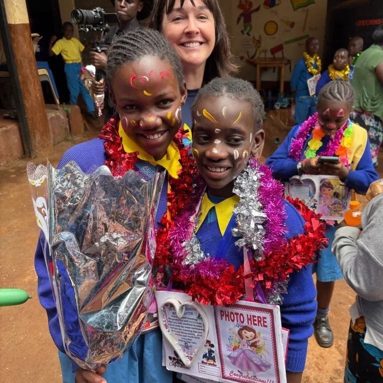 Two smiling young girls with face paint and decorations around their necks, standing close together, holding a bouquet and a photo frame with a cartoon princess on it. A woman stands behind them, also smiling, with other children and adults in a colo