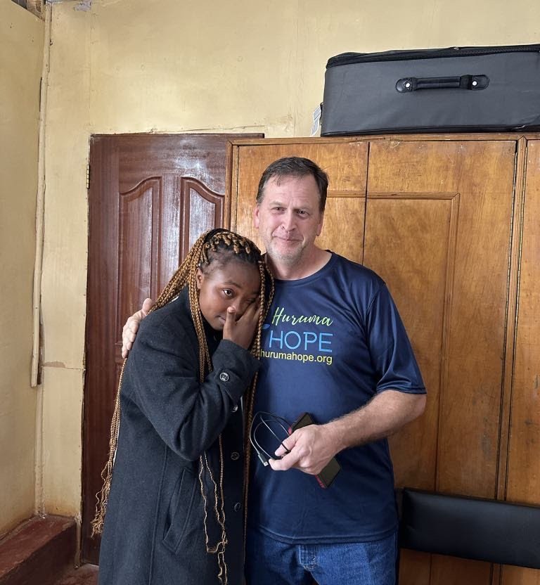 A man and a woman standing close together indoors. The woman covers her face with her hand. The man wears a blue shirt with 'Huruma HOPE' written on it. The background shows wooden cabinets and a black suitcase on top of the cabinet.
