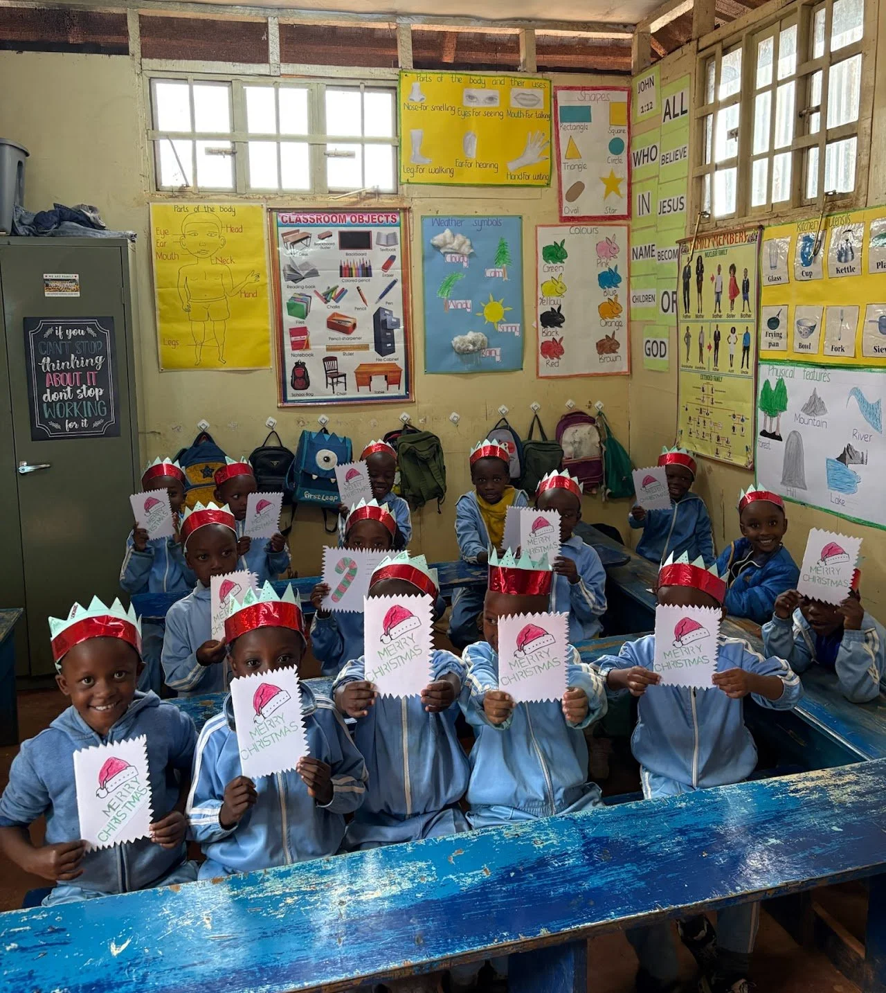 A classroom filled with children wearing Christmas hats and holding Christmas cards, smiling at the camera.