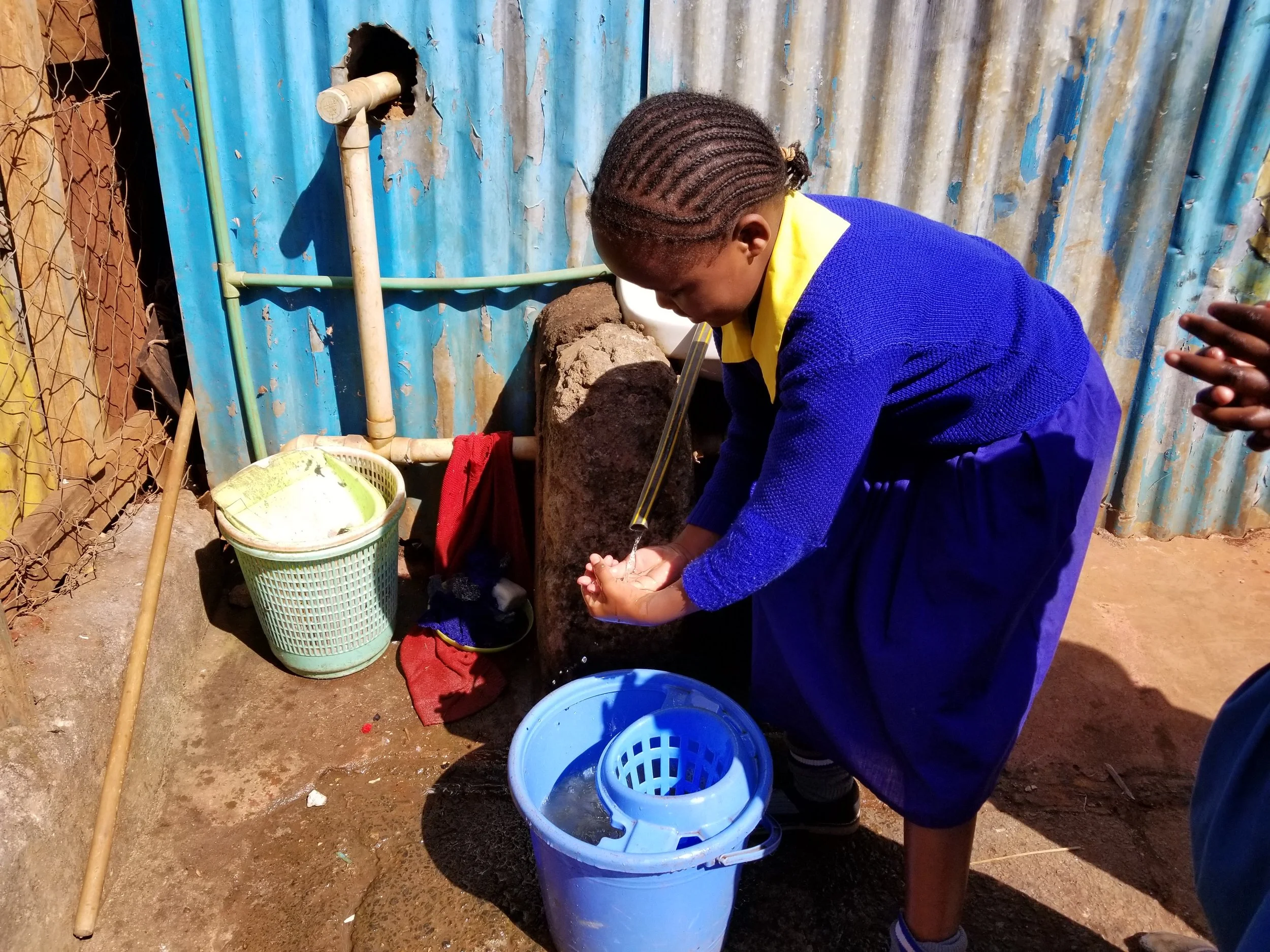 Girl in blue school uniform washing her hands at outdoor handwashing station with a blue bucket, water, and soap, set against a corrugated metal wall.