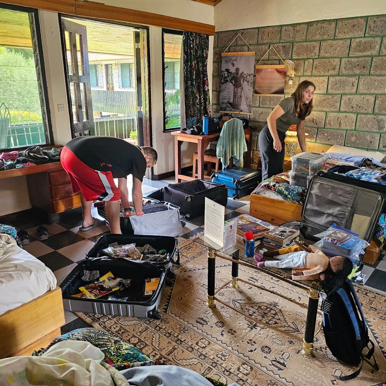 Two people packing luggage in a room with open door and windows, cluttered suitcases, a bed, a table, and various personal items.