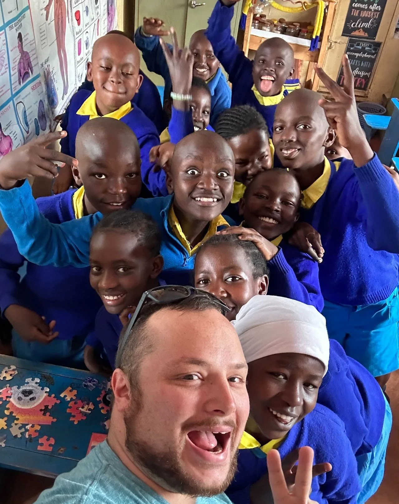 A group of smiling children and an adult taking a selfie in a classroom. The children are wearing blue and yellow uniforms, and some are making peace signs or excited gestures. The classroom has educational posters and colorful decorations.
