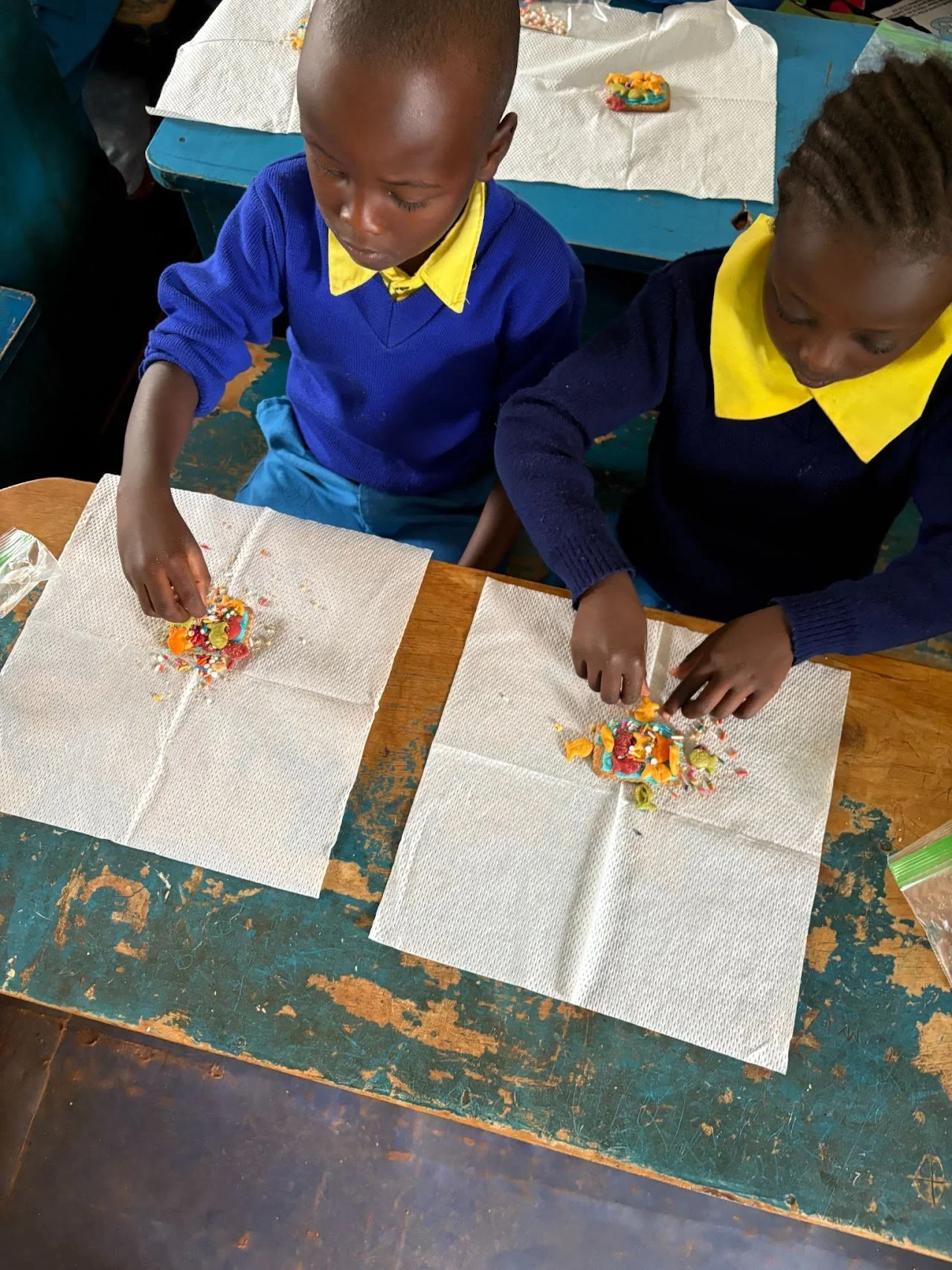 Two children in school uniforms decorating cookies with colorful sprinkles at a wooden table.