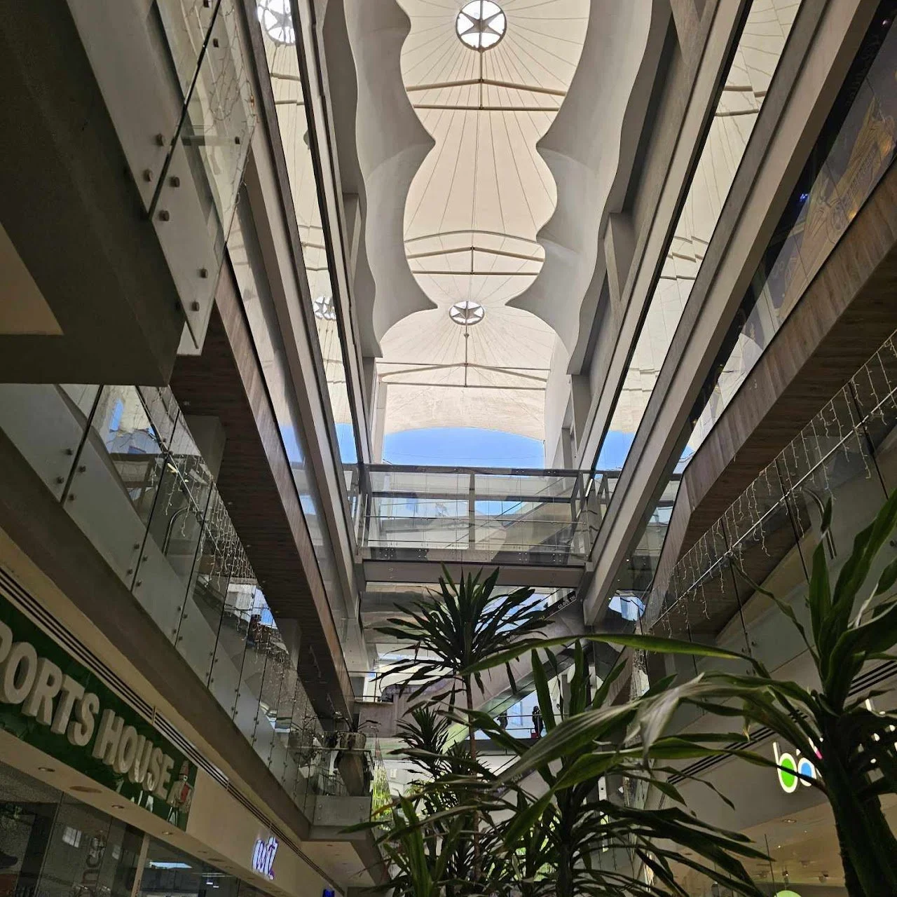 Interior view of a multi-story shopping mall with a glass ceiling and decorative overhead structure, plants in the foreground, and shops on the lower level.