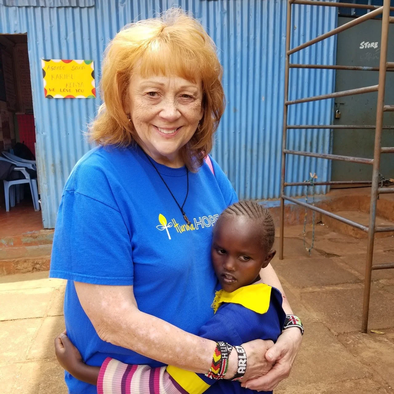 A woman with orange hair and a blue T-shirt is hugging a young girl with braided hair and a yellow and blue school uniform outside near blue corrugated metal walls.