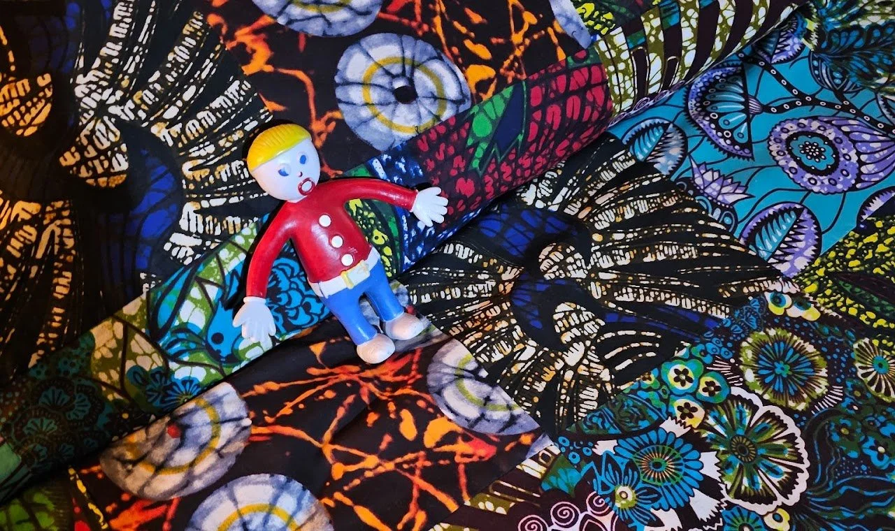 A small plastic figurine of a boy with blond hair, wearing a red shirt with white buttons, blue pants, and white shoes, lying on a colorful, patterned fabric with vibrant butterfly and floral designs.