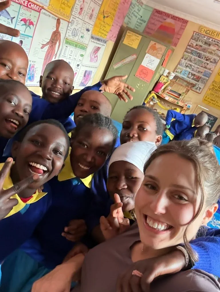 Group of smiling students and a woman taking a selfie in a classroom with educational posters on the wall.