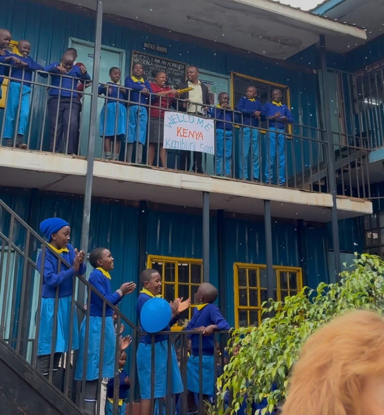 Children in school uniforms on outdoor staircases and balcony, celebrating with applause and a sign that says 'Welcome Kenya Karibu Sano' during a school event in Kenya.