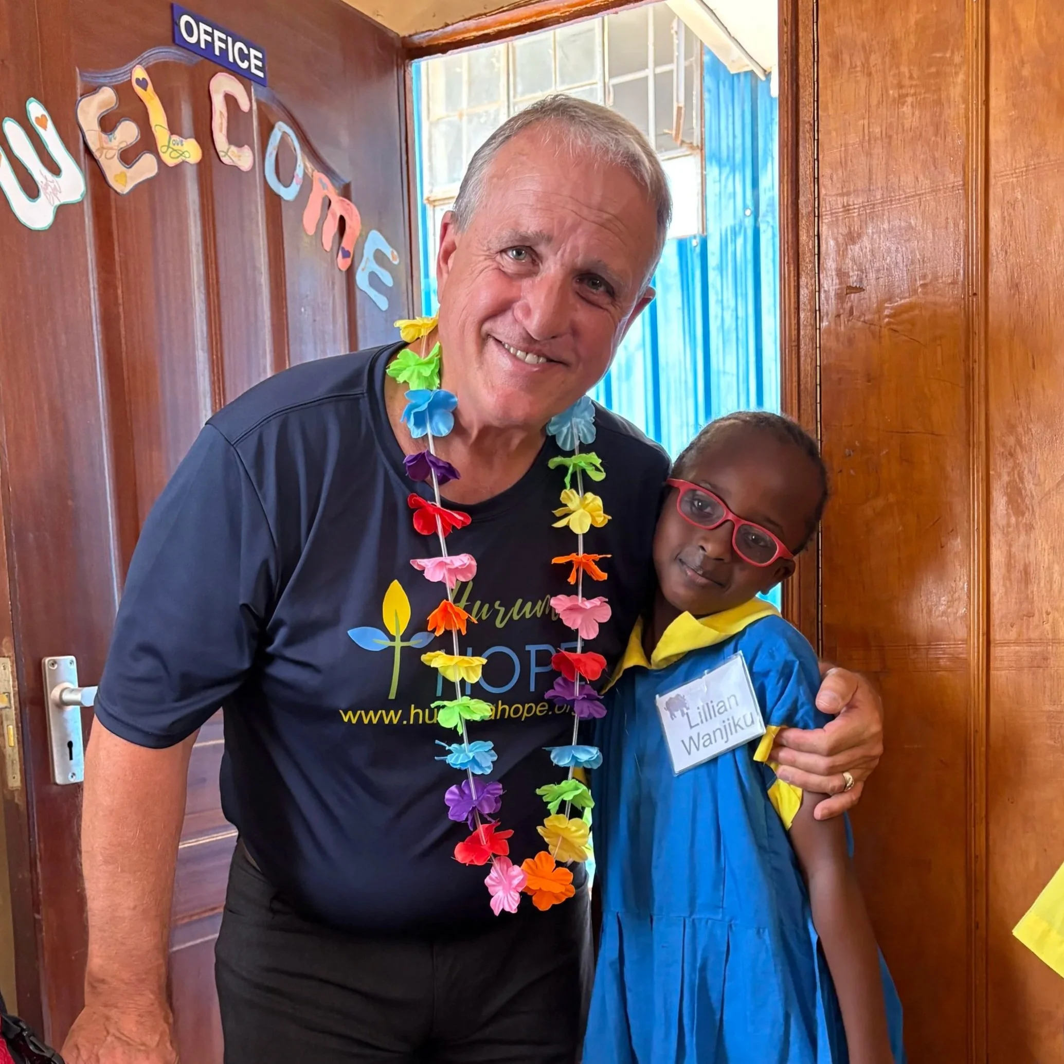 A man and a young girl smiling and hugging indoors. The man is wearing a colorful flower lei and a black T-shirt with a logo; the girl is wearing glasses and a blue uniform with a name tag that reads "Lillian Wanjiku." There are decorative letters in the background, and the setting appears joyful and celebratory.