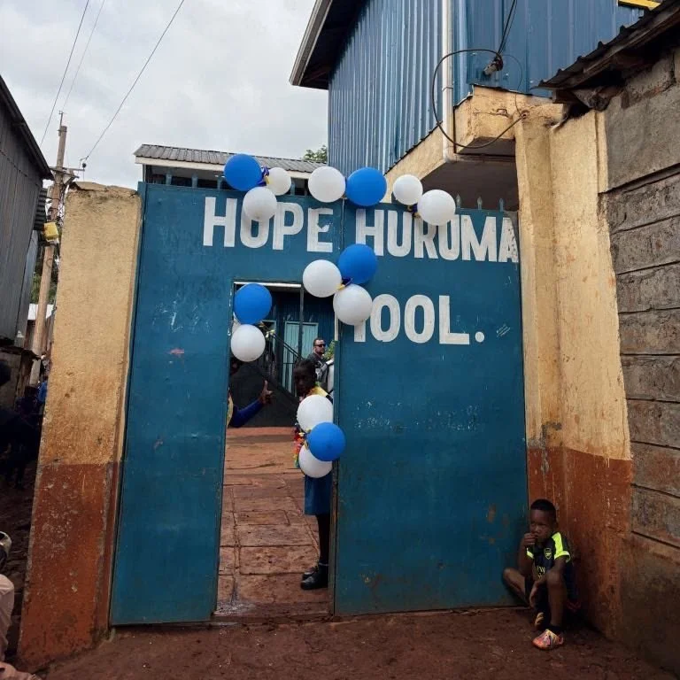 Blue gate with white and blue balloons and the words 'Hope Huruma Pool' painted above. Children are visible inside and outside the gate in an outdoor area with buildings.