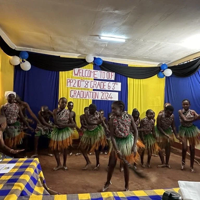 Group of young girls dressed in colorful tribal costumes dancing at graduation celebration, decorated with blue, yellow, and black drapes, balloons, and a sign that reads 'Welcome to our PP2 10th & Grade 6 3rd Graduation 2024'.