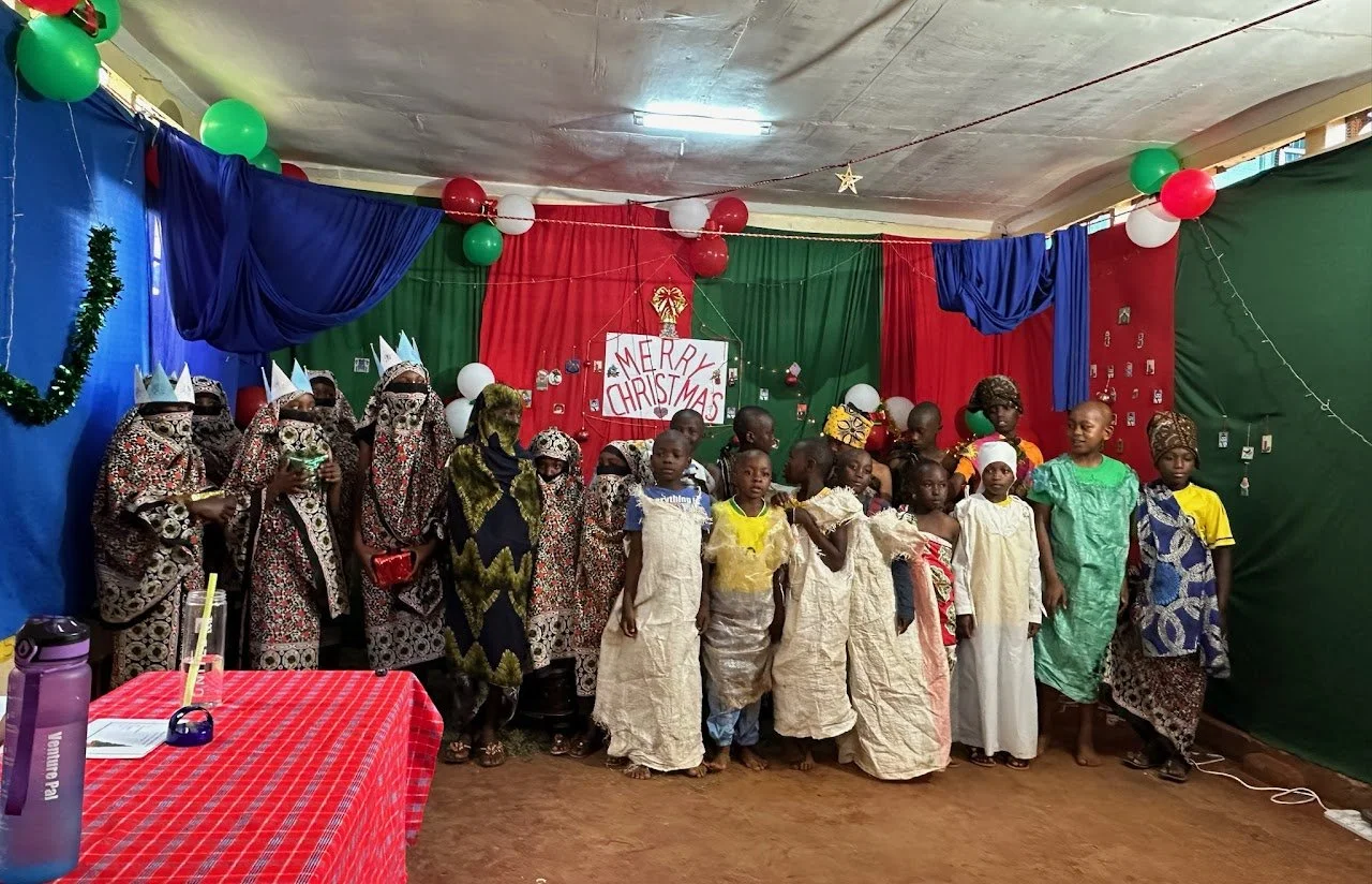 Children and adults in festive attire standing in front of a decorated background with a 'Merry Christmas' sign, Christmas ornaments, and festive decorations.