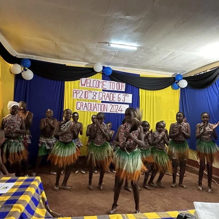 Group of children dancing in traditional attire on stage at a grade 6 graduation ceremony with a welcome banner and colorful decorations in the background.