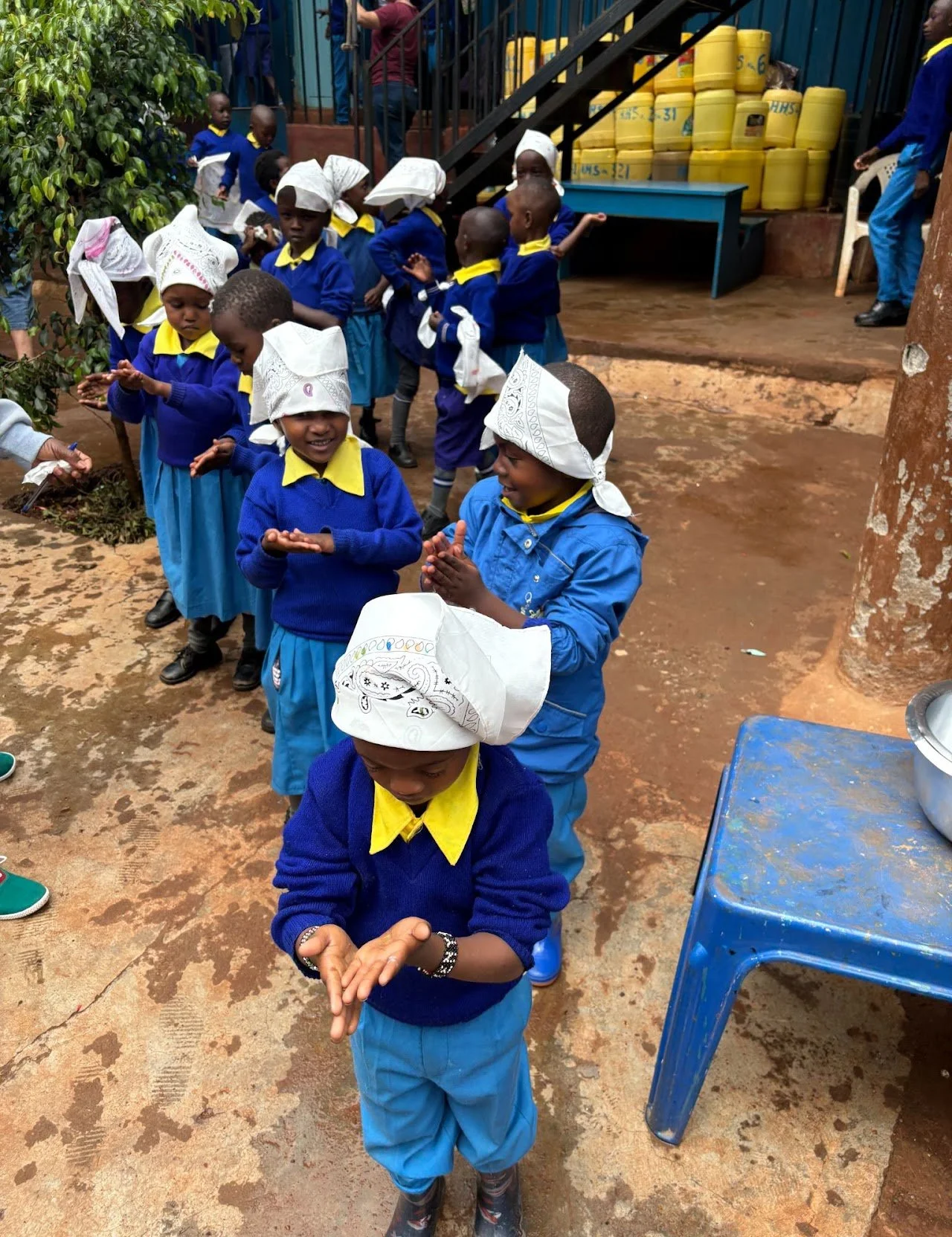 Children wearing blue uniforms and white headscarves, standing in line outdoors, some clapping and smiling, with a blue table and yellow containers in the background.