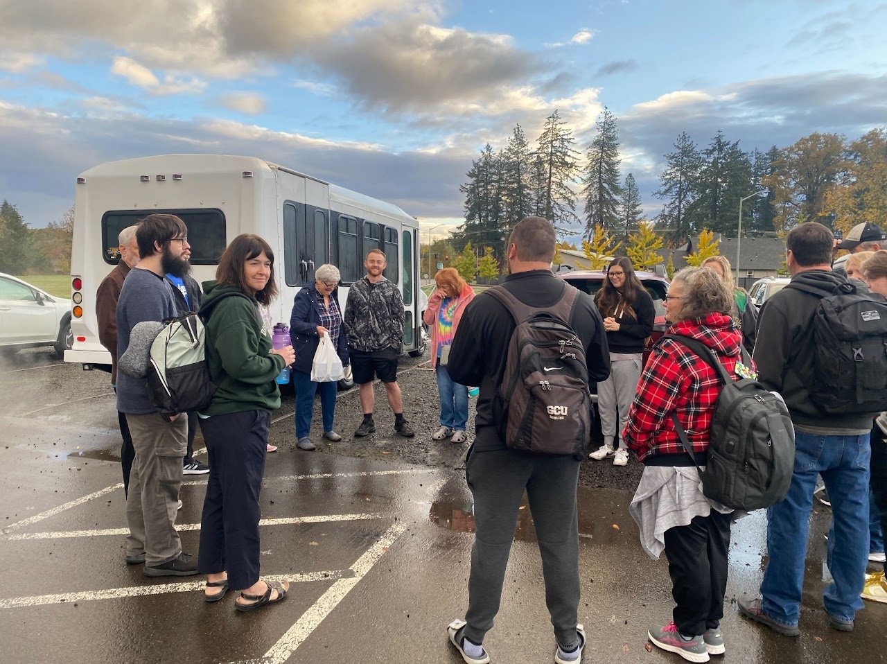 Group of people gathered in a parking lot near a large white bus, some holding backpacks and water bottles, with a cloudy sky and trees in the background.