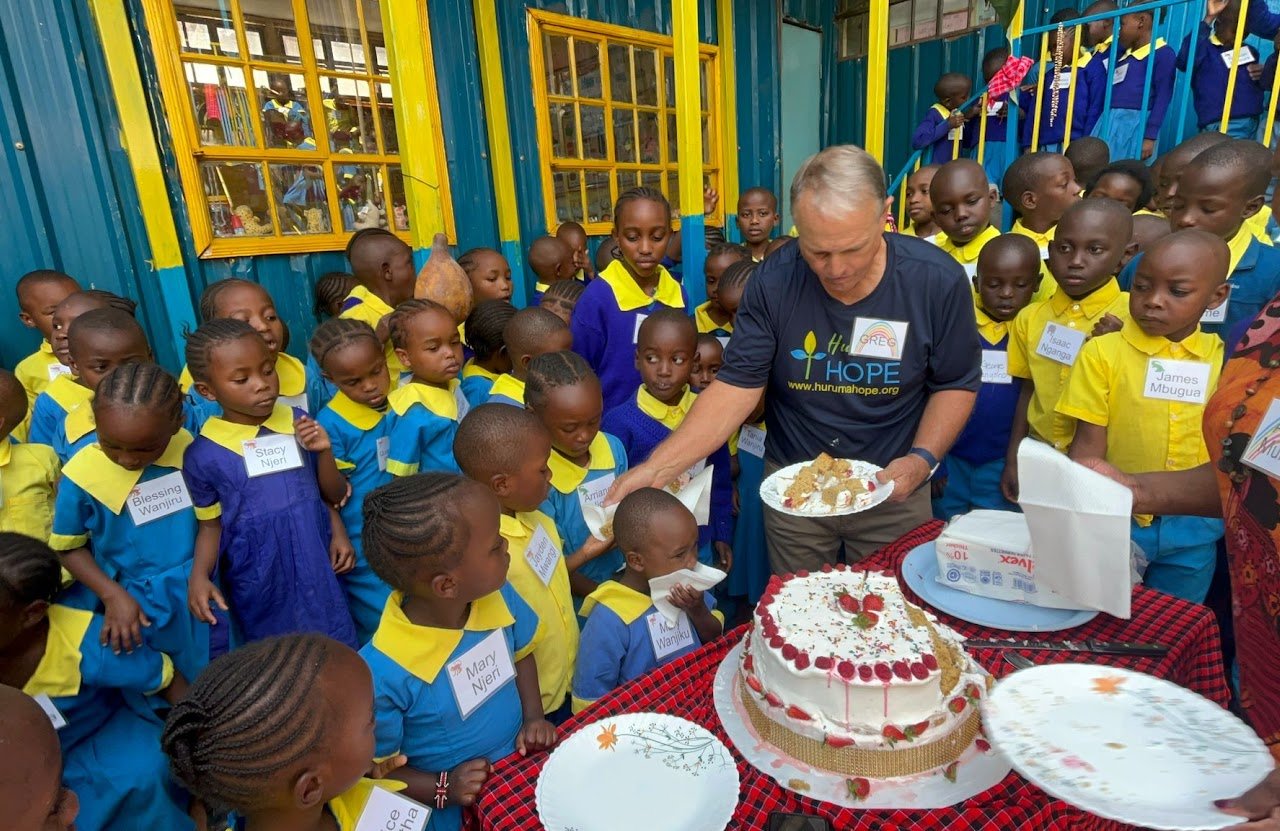 A group of children in school uniforms gathered around a man who is serving cake at a celebration outdoors. The children are wearing yellow and blue uniforms and are watching the cake being served. The setting appears to be at a school with colorful 