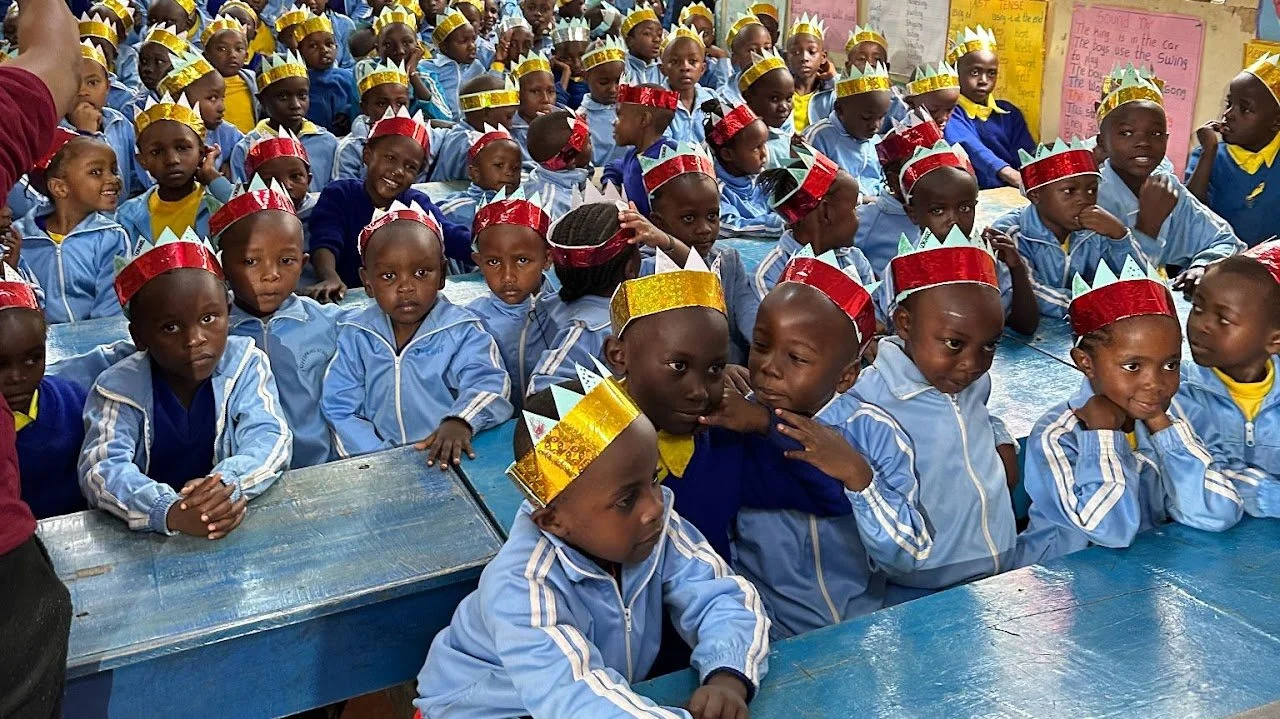 Group of young children in school uniforms with crowns, sitting at desks in a classroom decorated with posters.
