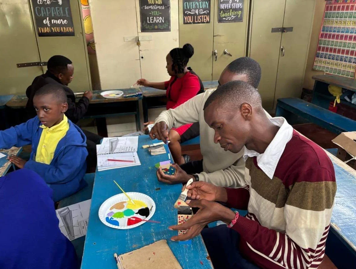 Adults and children in a classroom painting and doing arts and crafts at desks with colorful posters and a calendar on the walls.