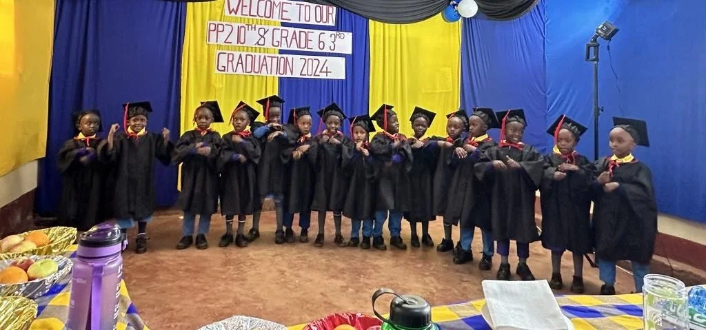 A group of young children in graduation caps and gowns standing on a stage at their graduation ceremony, with a banner above them that says 'Welcome to our PP2 10th & Grade 6, 3rd Graduation 2024' and a decorated background with yellow, blue, and bla