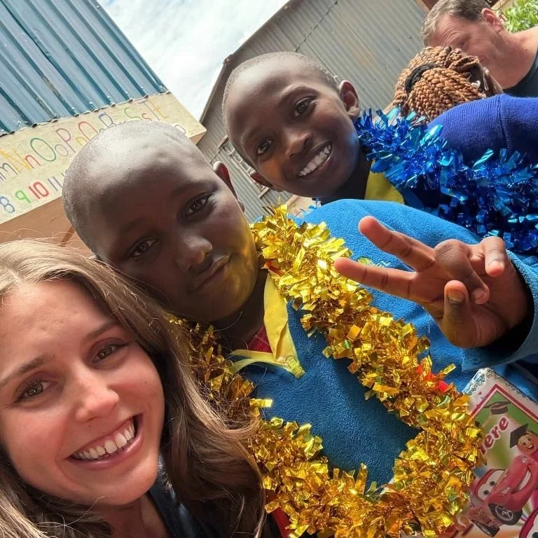 Group of children and a woman smiling in a festive setting, with children wearing gold and blue tinsel necklaces, one making a peace sign, in an outdoor classroom.