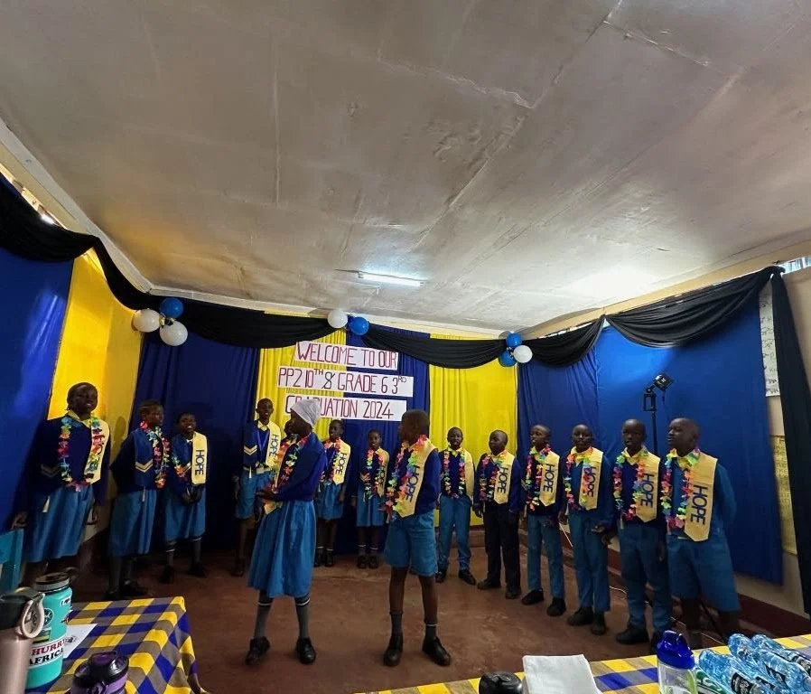 A group of children in blue uniforms and sashes that say 'HOPE' standing on stage during a graduation ceremony, with a colorful banner that reads 'Welcome to our PP2 2nd & 3rd Grade Graduation 2024' in the background, decorated with black, yellow, an