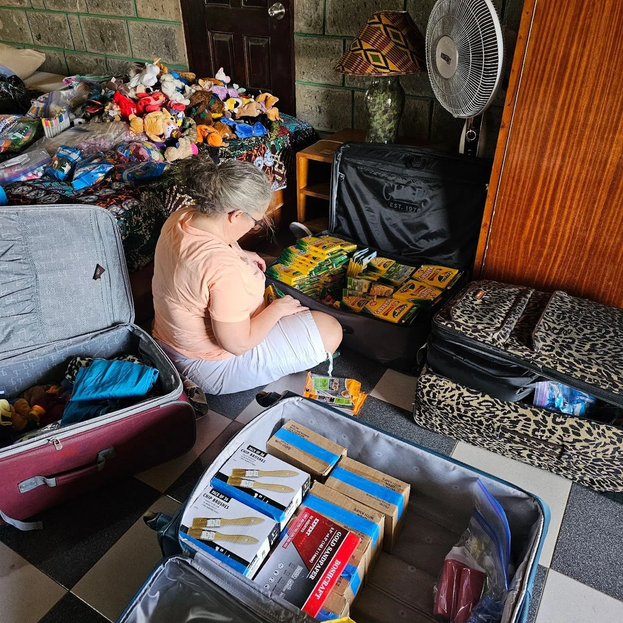 A woman is sitting on the floor, packing a suitcase with boxes of gardening tools and supplies in a room filled with stuffed animals, toys, and other packed luggage.