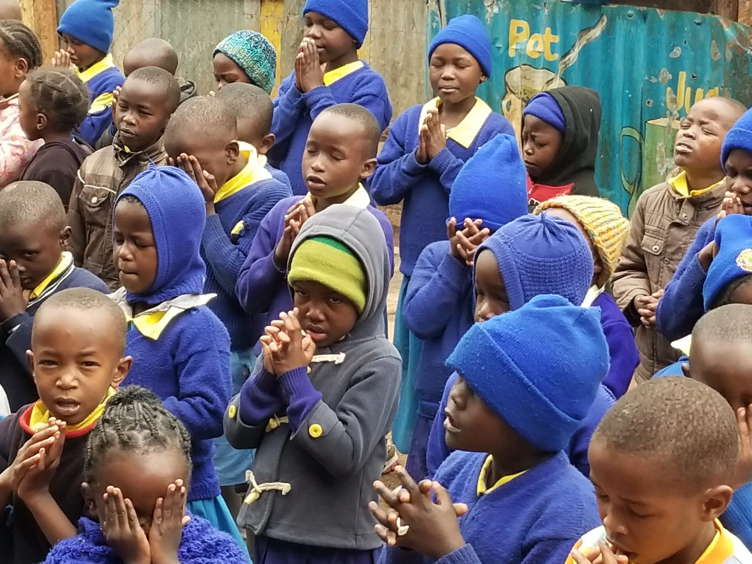 Group of children praying outdoors, some with eyes closed and hands clasped, wearing warm clothing.